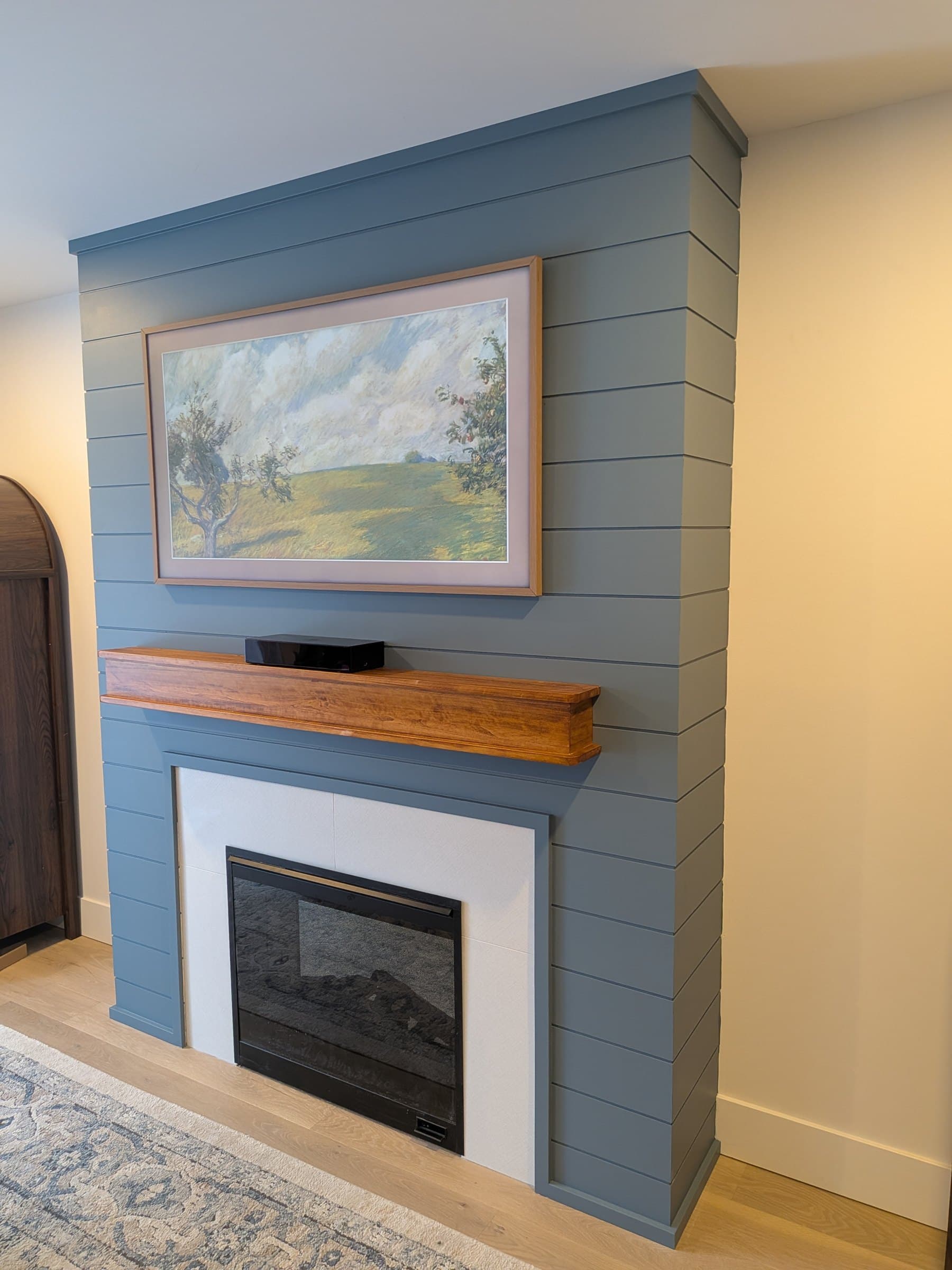 Basement fireplace with a blue panelled feature wall, wood mantel, and framed artwork